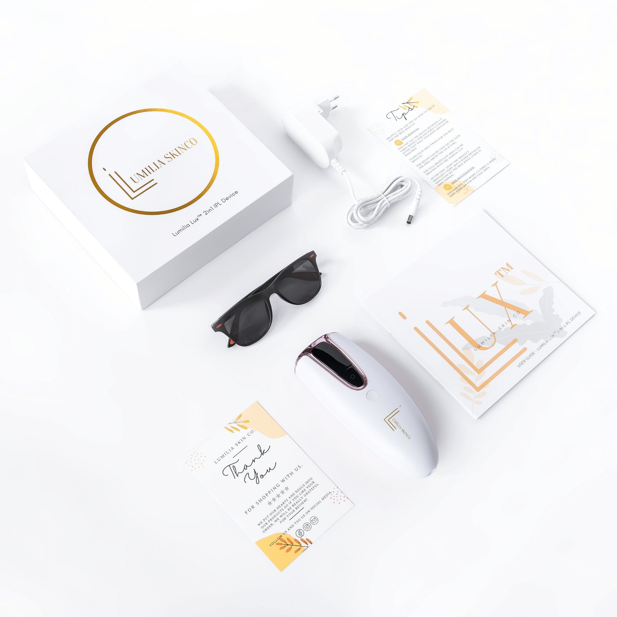 White Lumilia Lux 2in1 IPL laser hair removal and skin rejuvenation handset with silver trim, laid out with full packaging: branded Lumilia Skin Co. box, charger, manual, sunglasses, and thank-you card.